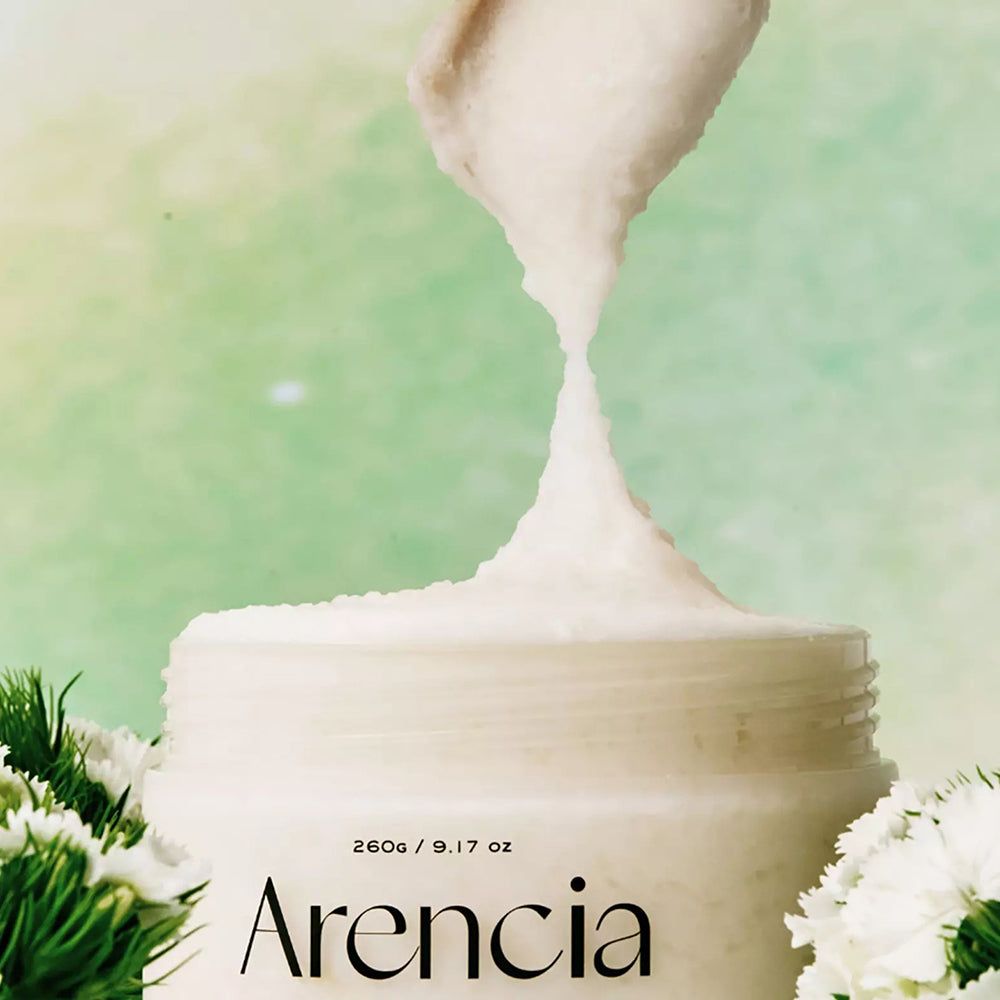 Scrub ARENCIA prelevato da un vasetto bianco. Texture leggera, circondato da fiori bianchi.