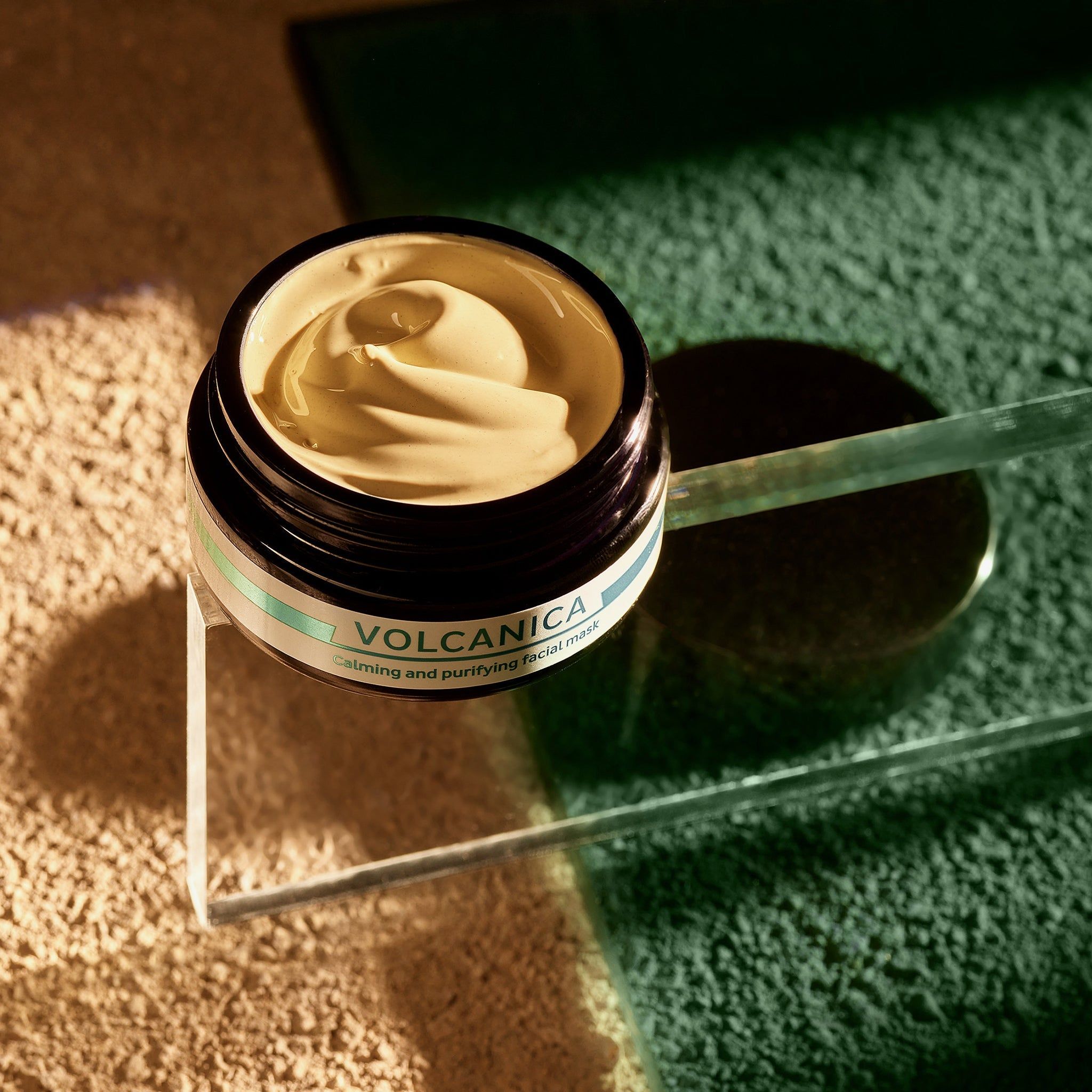 Vasetto aperto con maschera cremosa. Scritta: VOLCANICA, Calming and purifying facial mask. Su lastra di vetro.