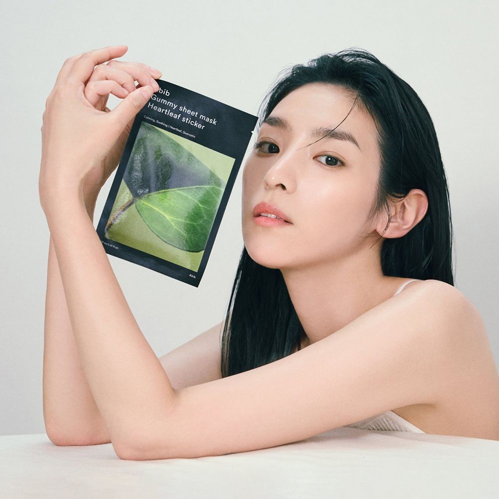 Donna che tiene in mano confezione nera con nome prodotto e motivo a foglia. Testo: ABIB Gummy Sheet Mask Heartleaf sticker.