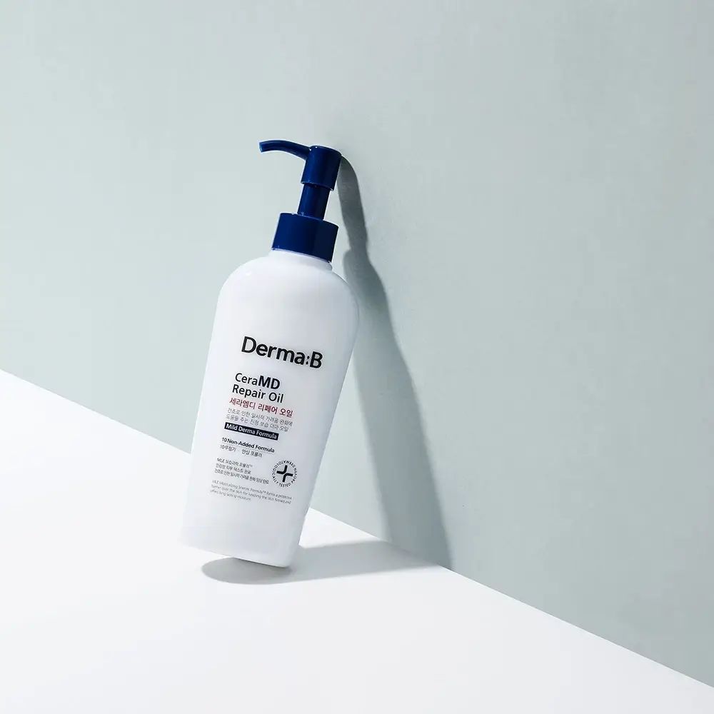 Flacone bianco con erogatore blu. Scritta Derma:B CeraMD Repair Oil. Prodotto appoggiato a parete azzurra.