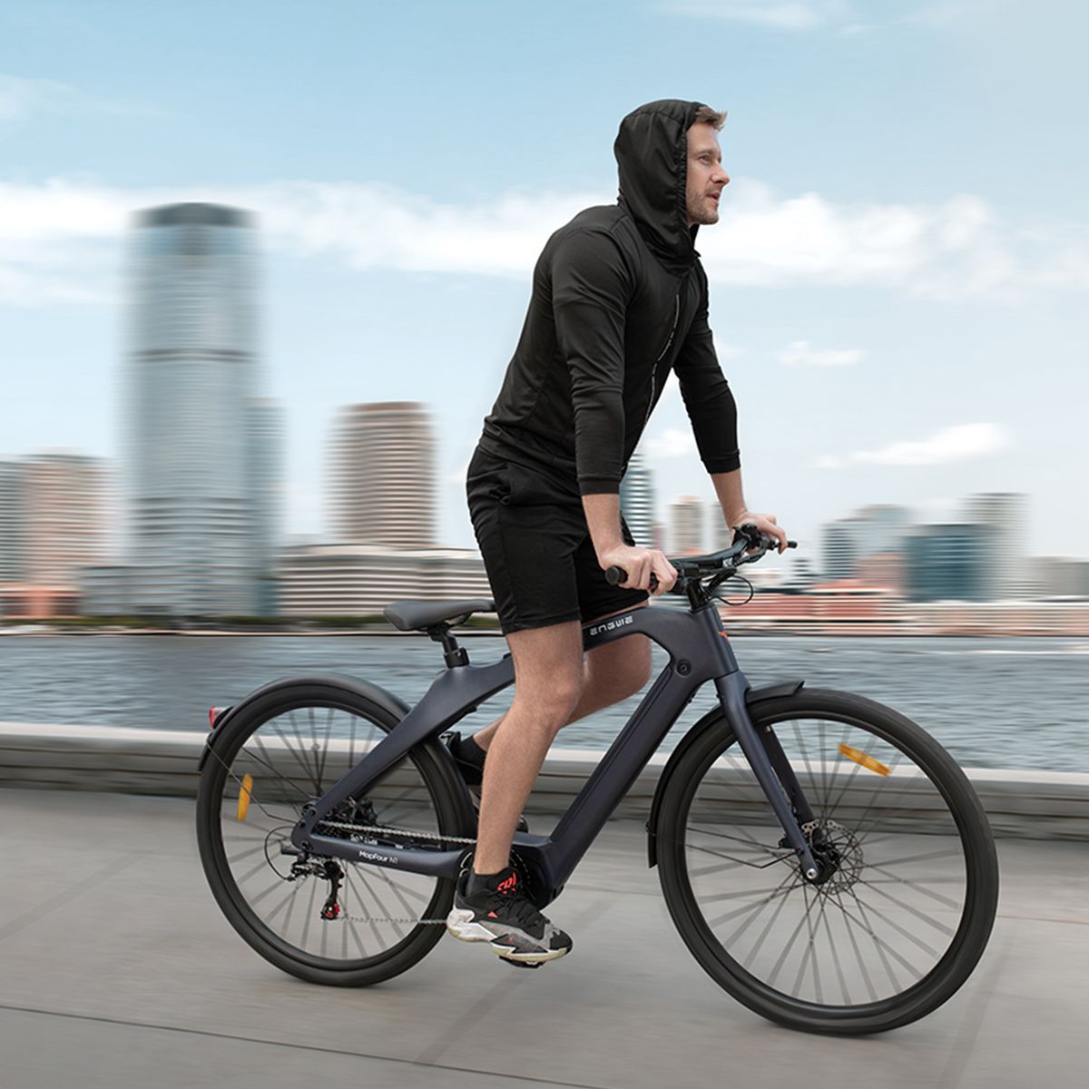 Persona in bicicletta elettrica grigio scuro. Paesaggio urbano sullo sfondo. Logo Engwe.