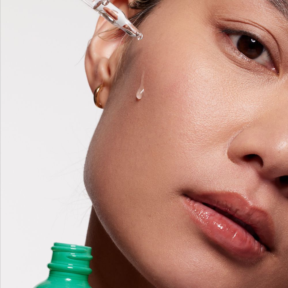 Primo piano di un viso. Una goccia di siero cade da una pipetta sulla pelle. Flacone verde sullo sfondo.