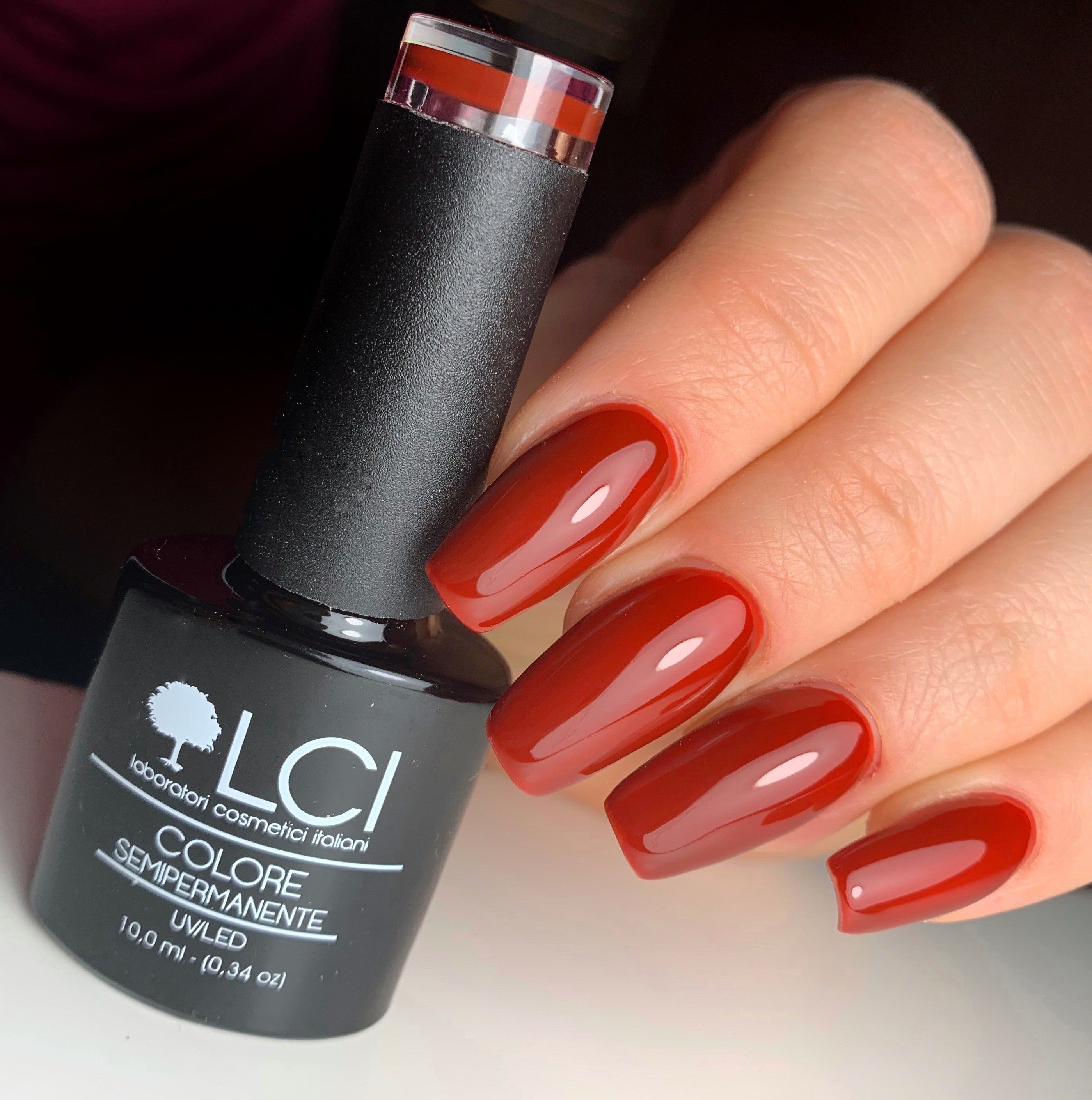 Mano con unghie smaltate e flacone di smalto. Unghie rosse e flacone di smalto nero. Scritta: LCI, Colore Semipermanente.