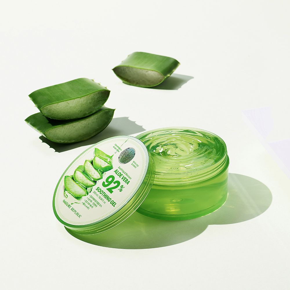 Gel verde in un contenitore trasparente con coperchio. Accanto, foglie di aloe vera. Sul coperchio, "Aloe Vera 92% Soothing Gel".