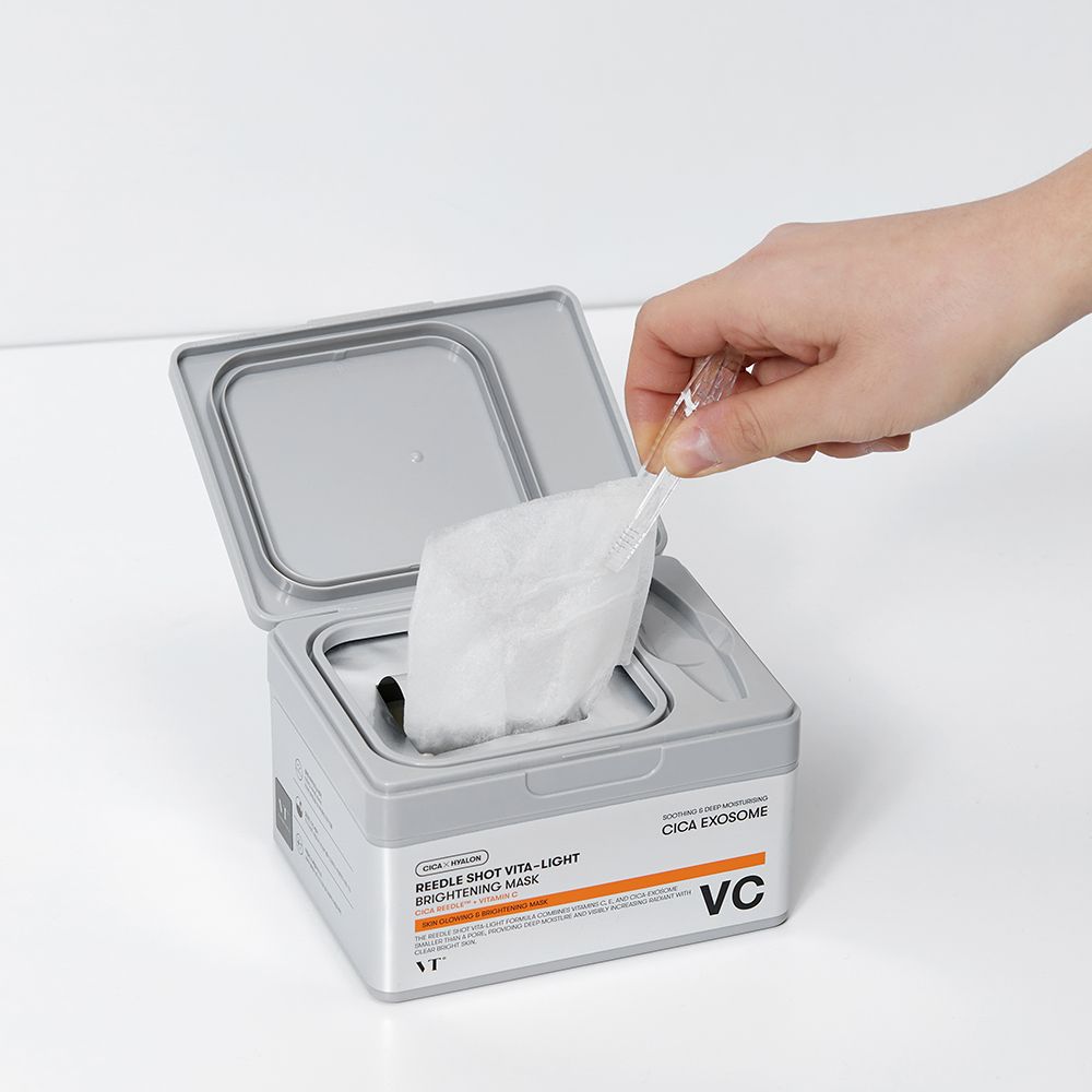 Mano che preleva una maschera da una scatola grigia. Scritta: Reedle Shot Vita-Light Brightening Mask. Marchio: VT. Confezione aperta.