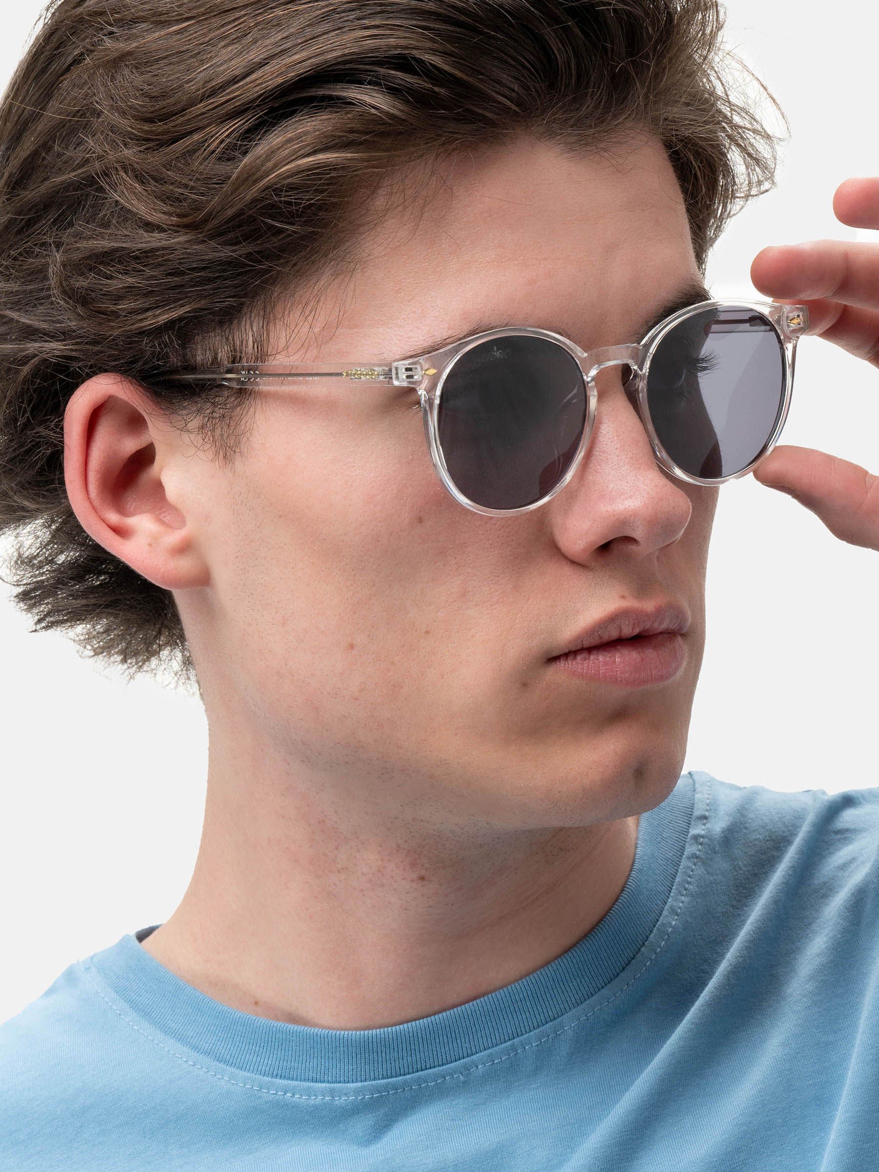 Persona che tiene occhiali da sole con montatura trasparente e lenti scure. Marchio: Smooder. Primo piano.