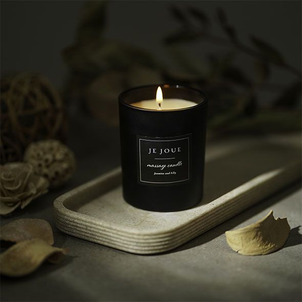 Candela nera accesa su un vassoio. Scritta: Je Joue, massage candle, Jasmine Lily. Decorazioni sullo sfondo.
