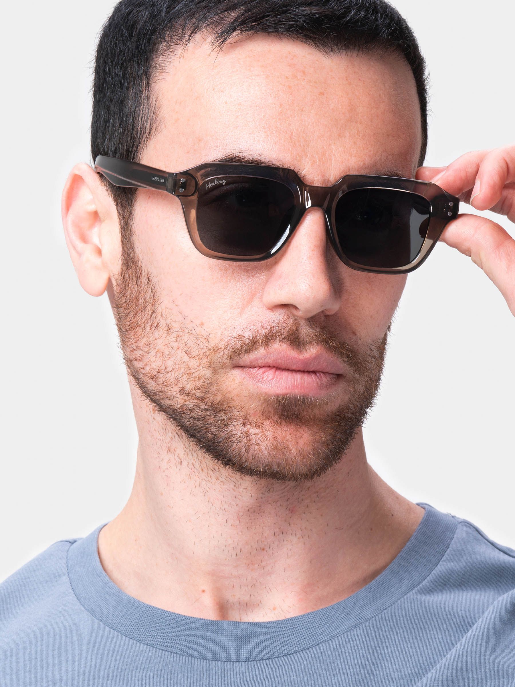 Uomo con occhiali da sole Soft Taupe Grey. Lenti rettangolari, montatura marrone chiaro. Logo Herling non visibile. Maglione blu.