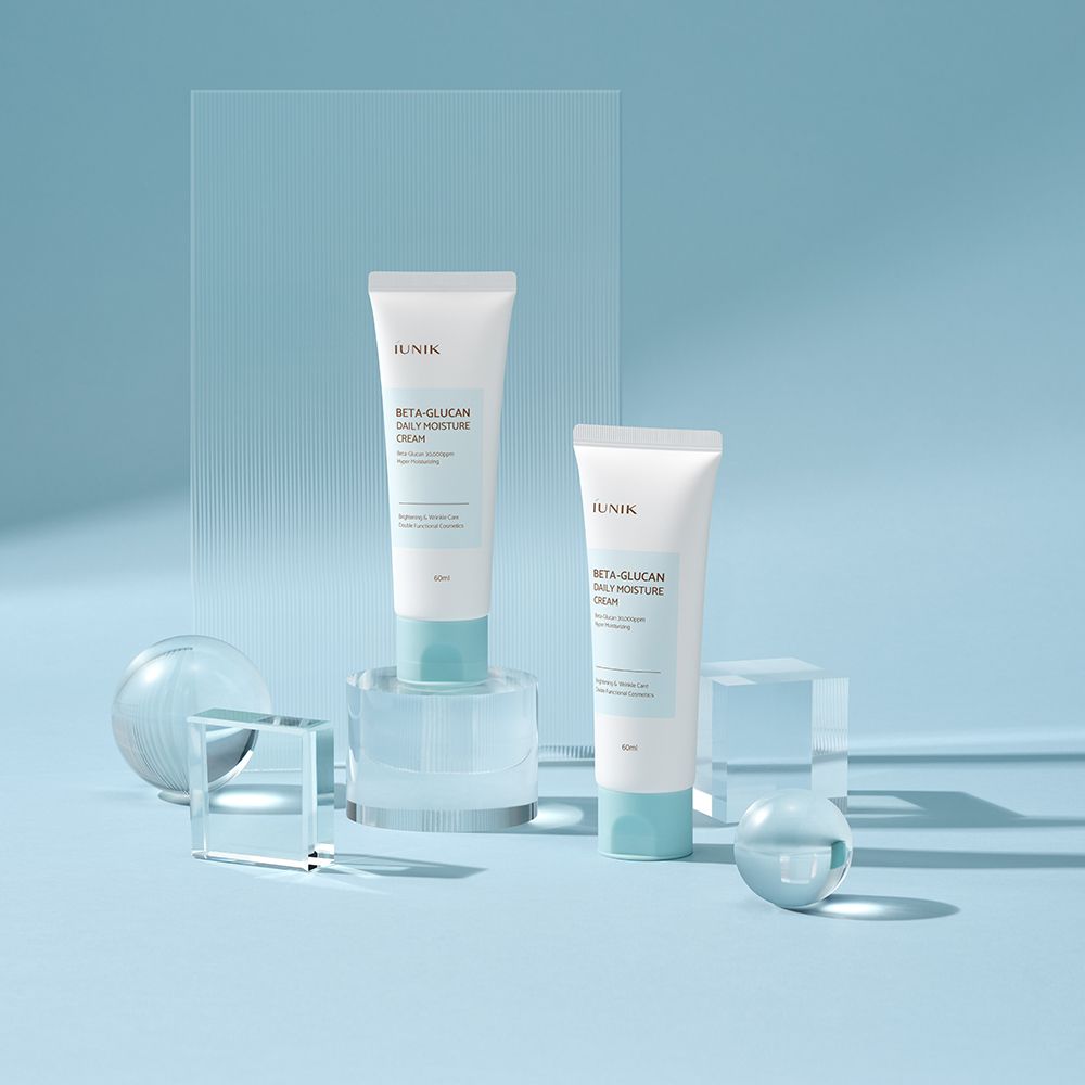 Due tubi di crema su piedistallo. Scritta: IUNIK, Beta-Glucan Daily Moisture Cream. Colori chiari.