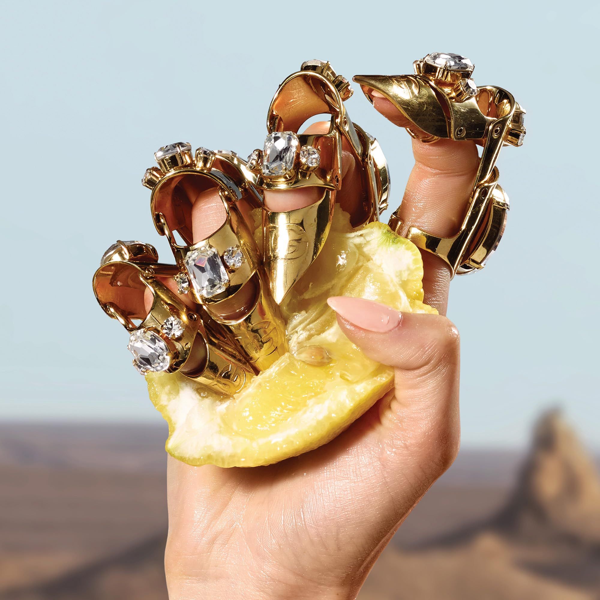 Mano con anelli dorati con pietre preziose. La mano tiene una metà di limone. Sfondo: deserto e cielo blu.
