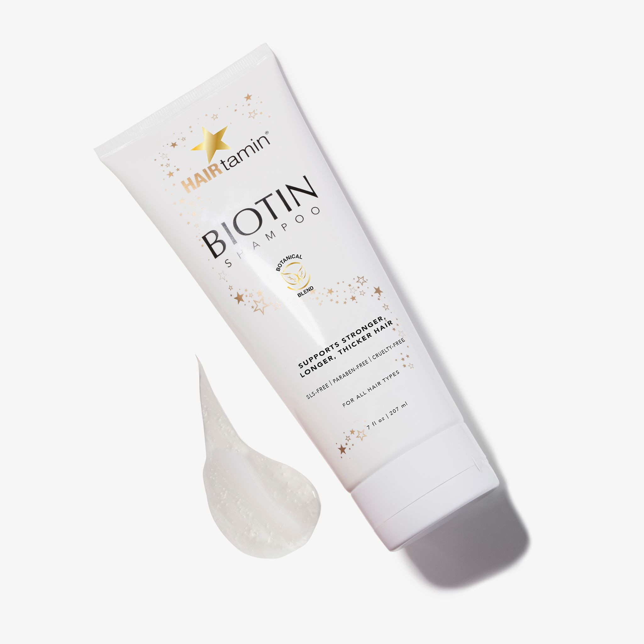 Tubo bianco "HAIRtamin Biotin Shampoo" con fuoriuscita. Stelle e scritta "Botanical Blend". Testo: "Supports stronger, longer, thicker hair".