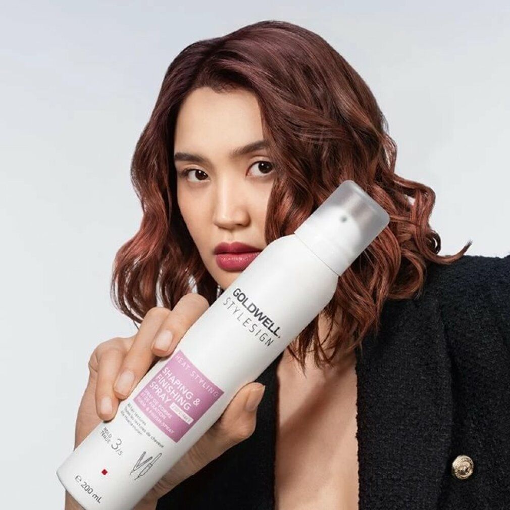 Una donna tiene in mano un flacone spray bianco. Scritta: Goldwell Stylesign Heat Styling Shaping & Finishing Spray. Indossa un blazer.