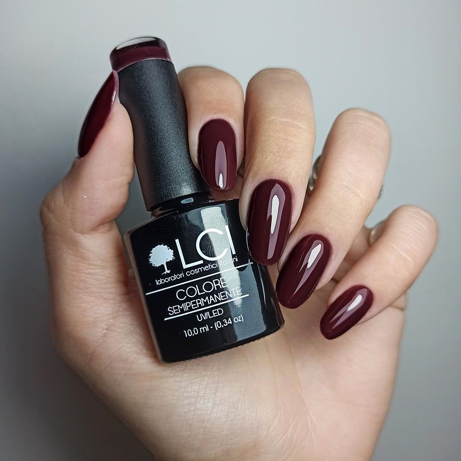 Mano con unghie smaltate e flacone di smalto. Colore: rosso-marrone. Scritta: LCI, Colore Semipermanente, UVLED 10,0 ml.