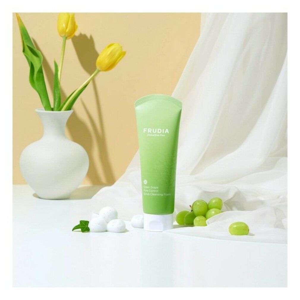 Tubo verde con tappo bianco. Scritta: FRUDIA. Testo: Green Grape Pore Control Scrub Cleansing Foam. Vicino a uva e vaso.
