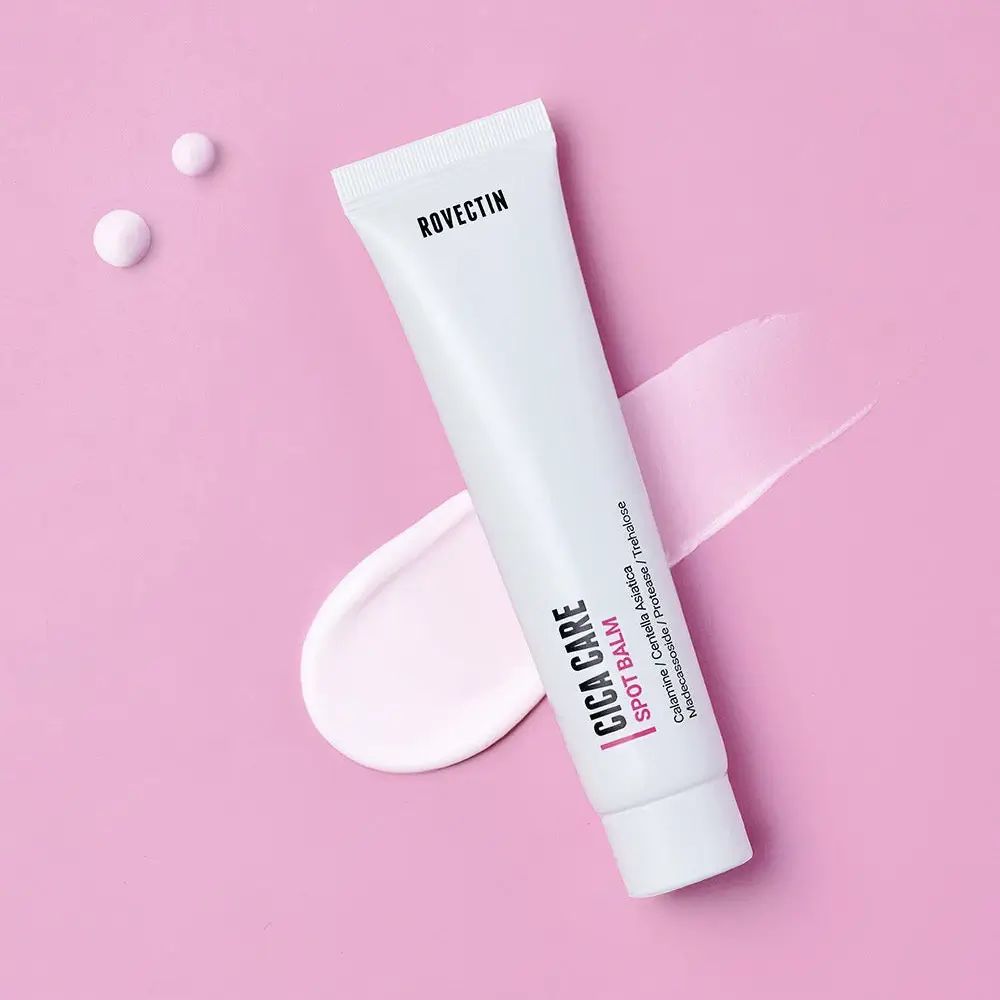 Tubo bianco con prodotto. Scritta: Rovectin, Cica Care Spot Balm. Sfondo rosa con sbavature di prodotto.