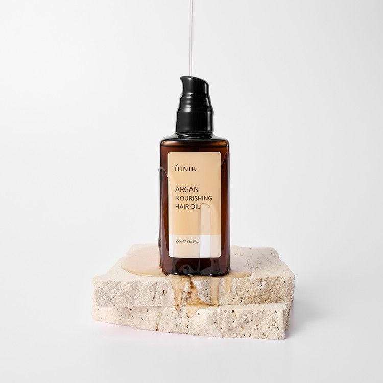 Flacone marrone con erogatore nero. Etichetta con nome del prodotto. Olio che gocciola su pietra. Scritta: IUNIK, Argan Nourishing Hair Oil.