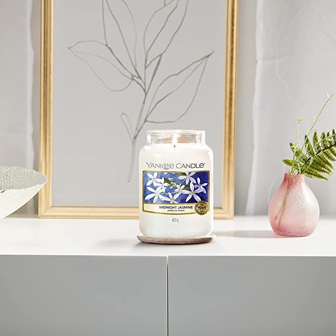 Candela profumata in vetro su un sottobicchiere. Adesivo con motivo floreale e scritta Midnight Jasmine. Sullo sfondo un'immagine.