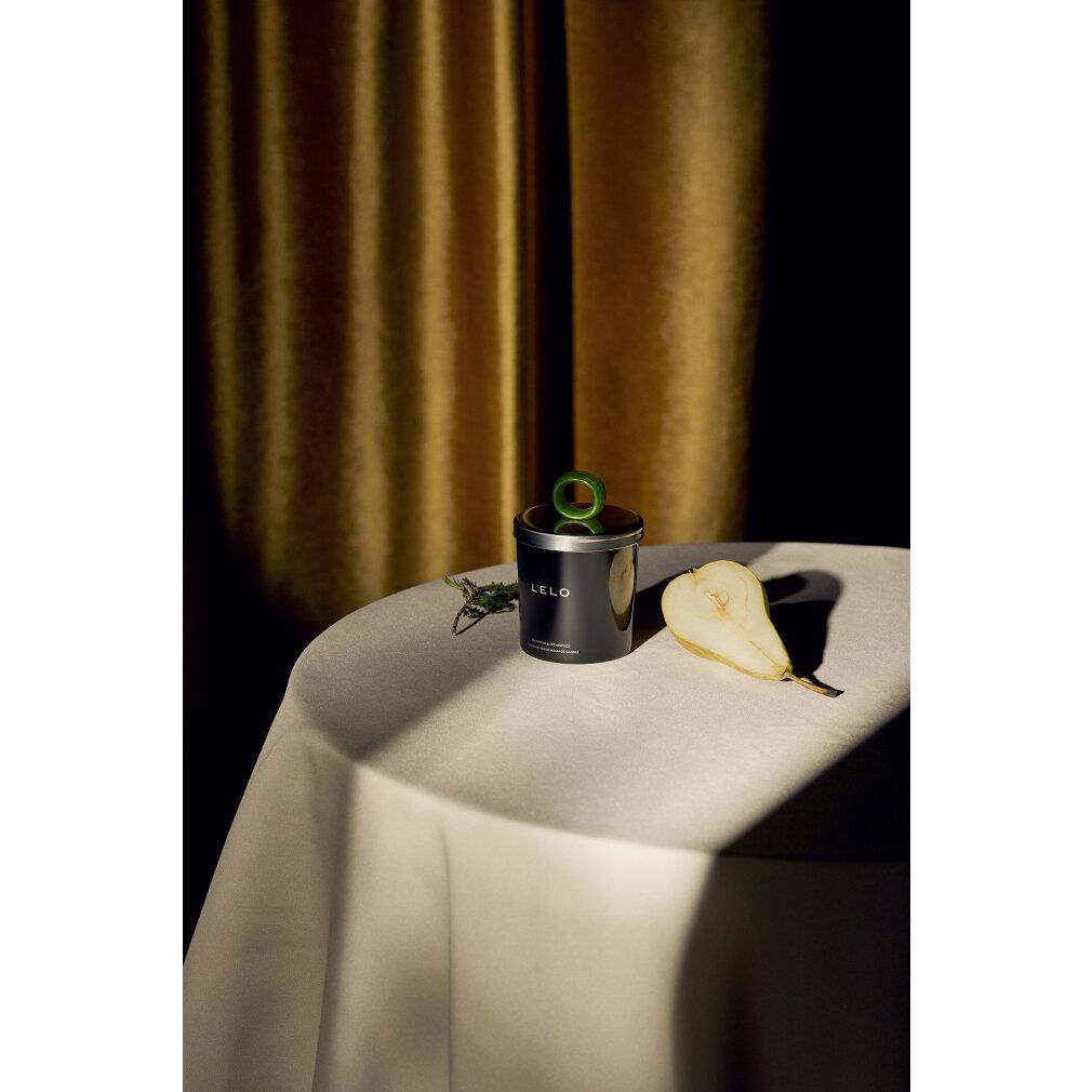 Barattolo nero con manico verde e pera su tavolo. Scritta LELO. Testo: Snow Pear & Cedarwood, FUCKING TOUCH MASSAGE CANDLE.