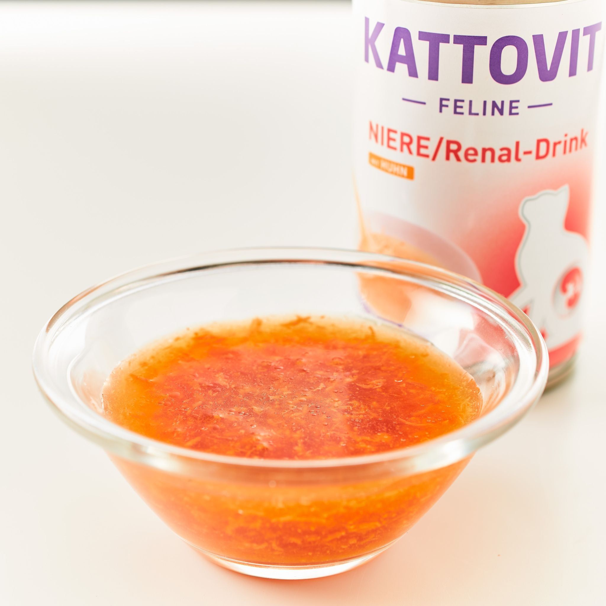Ciotola di vetro con liquido arancione. Sullo sfondo, una lattina KATTOVIT Feline Niere/Renal-Drink. Nome del prodotto e sagoma di gatto visibili.