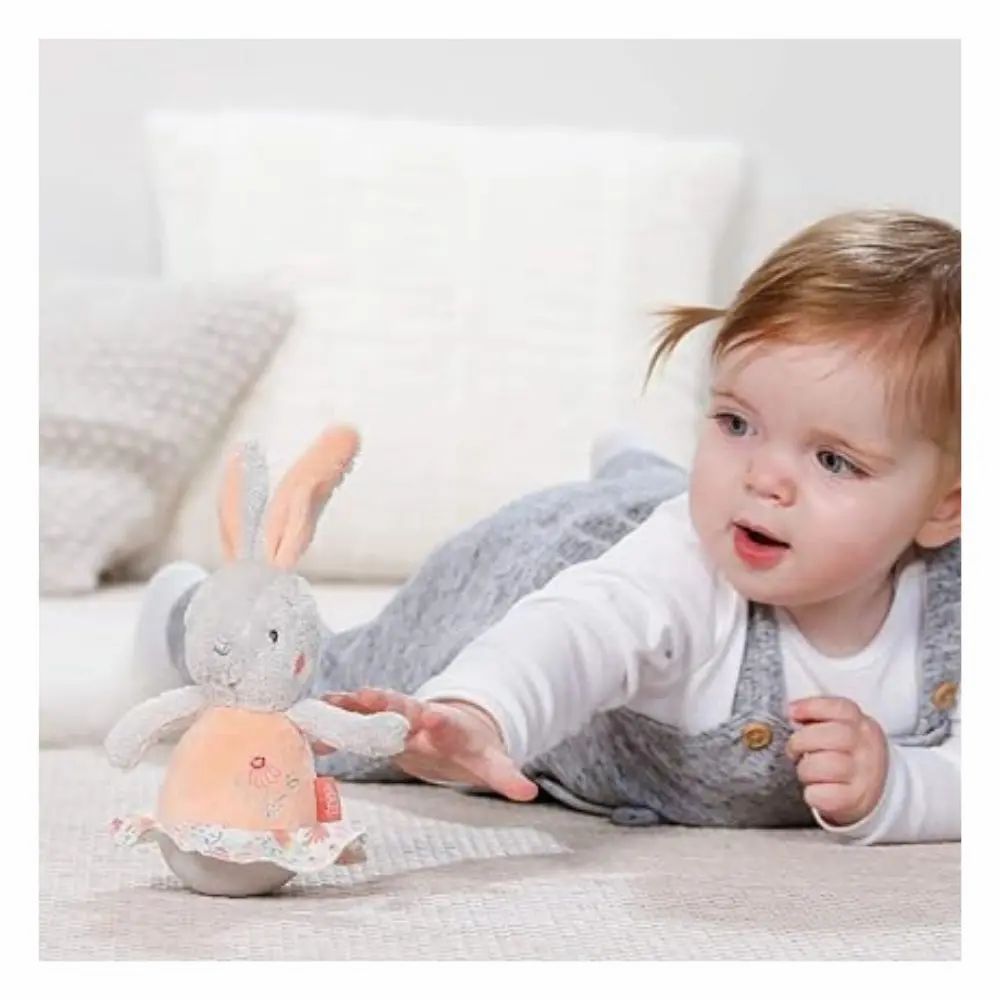 Bambino che afferra un coniglietto di peluche grigio, pancia e orecchie rosa. Sdraiato sul pavimento. Marchio: Fehn.