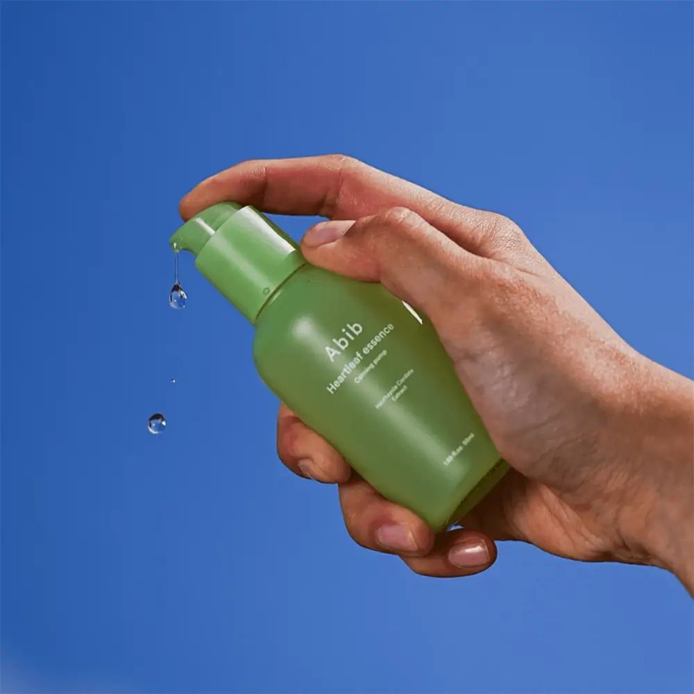 Flacone verde con pompa, tenuto in mano. Gocce che cadono dalla pompa. Scritta: Abib Heartleaf essence.