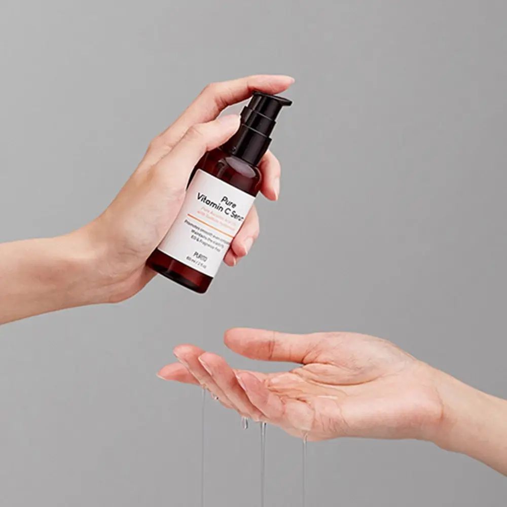 Mani che tengono un flacone di siero. Liquido che gocciola in una mano aperta. Scritta: Pure Vitamin C Serum.