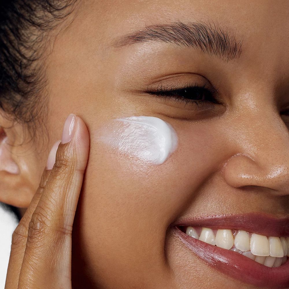 Persona applica crema sulla guancia. Crema bianca sulla pelle. La persona sorride.