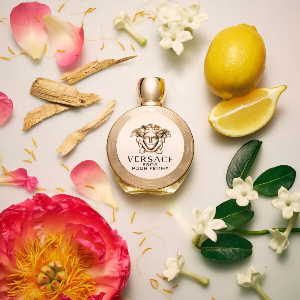 Flacone rotondo con logo Versace e scritta « Eros Pour Femme », circondato da petali, limone e legno.