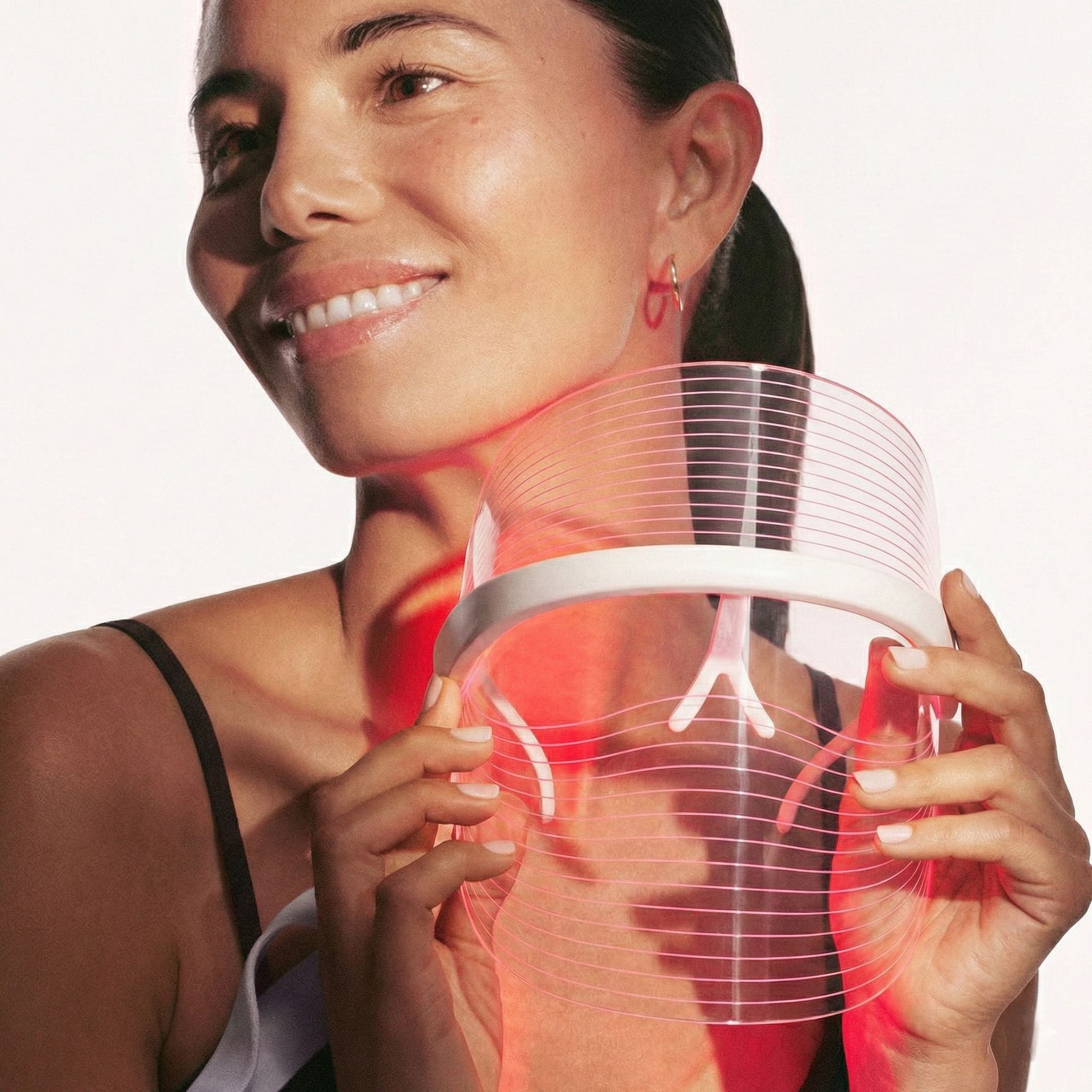 Una donna tiene in mano una maschera facciale LED bianca e trasparente, con luci rosse dirette verso la zona del collo.