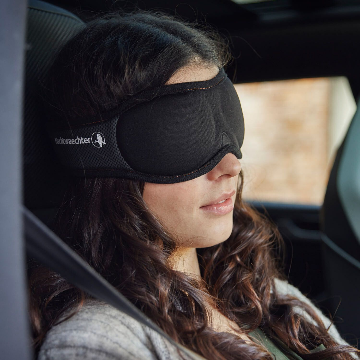 Donna in auto con maschera per dormire nera. Fascia grigia con logo, maschera sugli occhi.