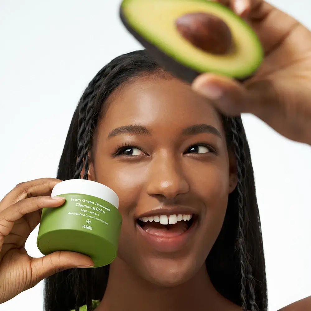Donna tiene vasetto verde e avocado. Scritta: From Green Avocado Cleansing Balm. Marchio: PURITO. La donna sorride.