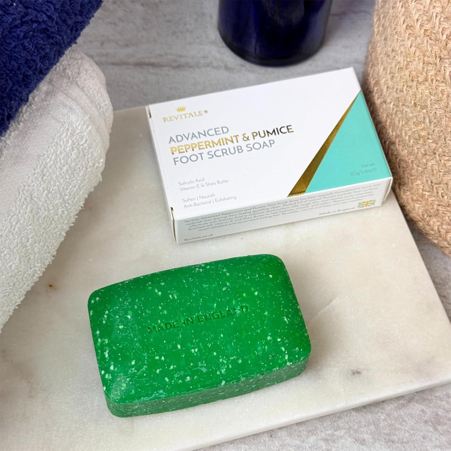 Sapone verde e confezione del prodotto su marmo. Asciugamano e cestino sullo sfondo.