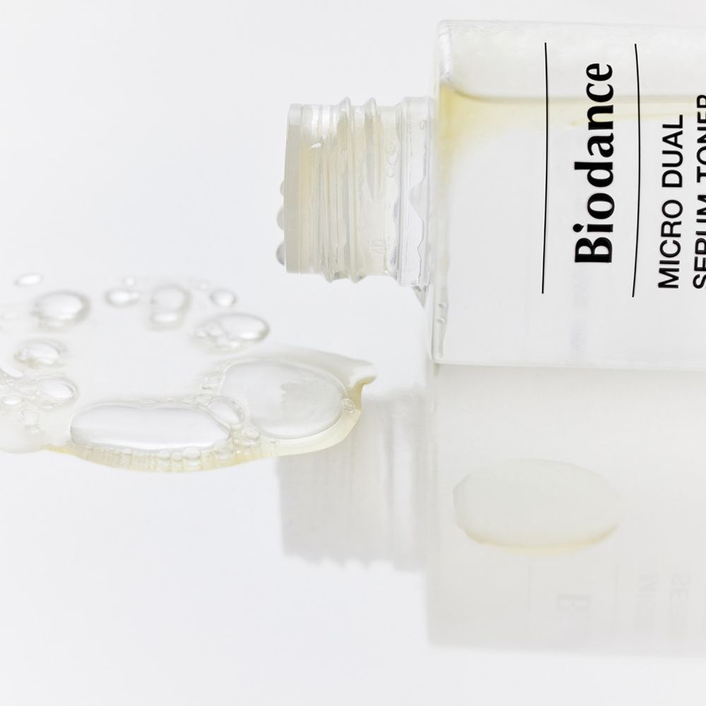 Flacone di BIODANCE Micro Dual Serum Toner appoggiato su una superficie. Il liquido fuoriesce dall'apertura. Liquido trasparente.