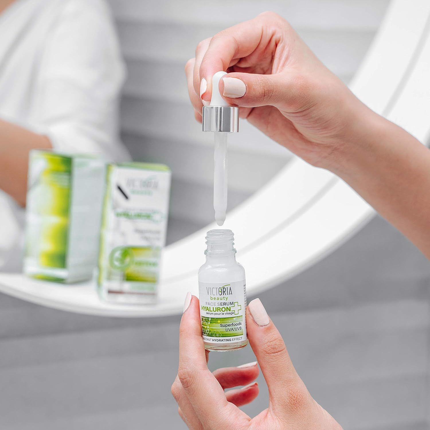 Flacone trasparente con contagocce. Scritta: Victoria Beauty Hyaluron Serum. Mani che tengono flacone e contagocce davanti allo specchio.