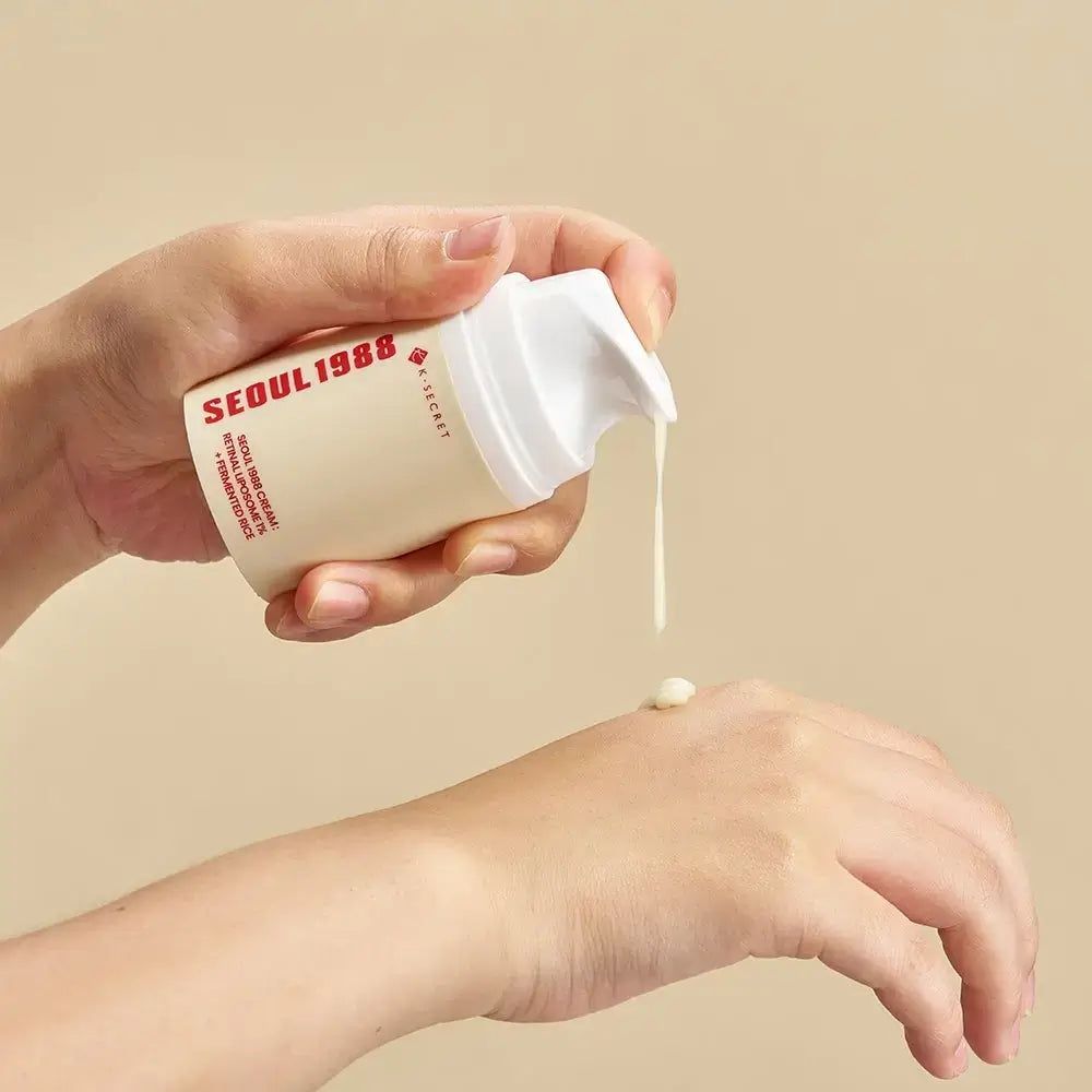 Persona che tiene un contenitore di crema e applica la crema sulla mano. Scritta: SEOUL 1988, K-SECRET. Testo: RETINAL LIPOSOME 1% + FERMENTED RICE.