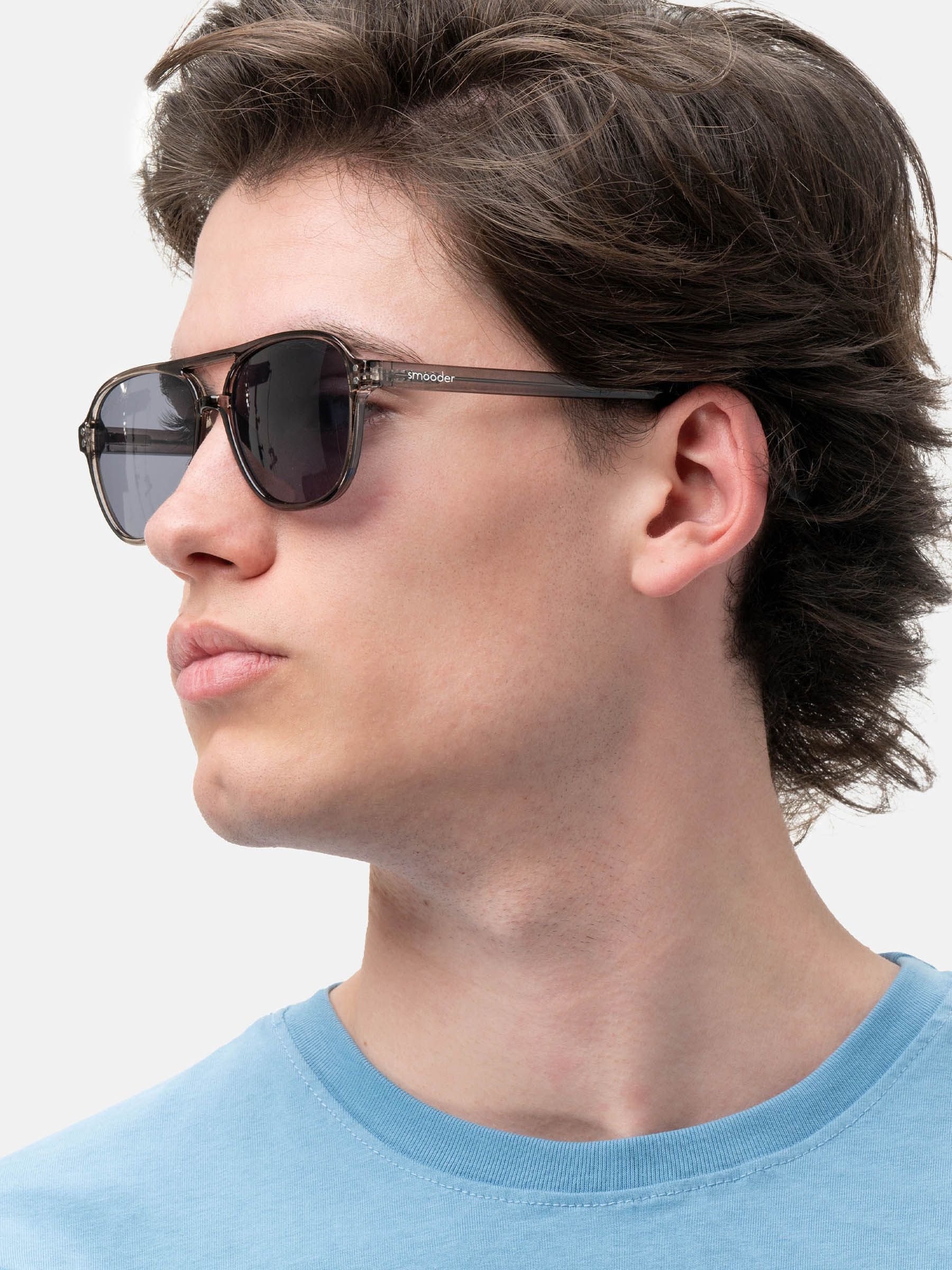 Persona con occhiali da sole grigi. Lenti scure, montatura trasparente. Marchio 'smooder' visibile. Maglietta azzurra.