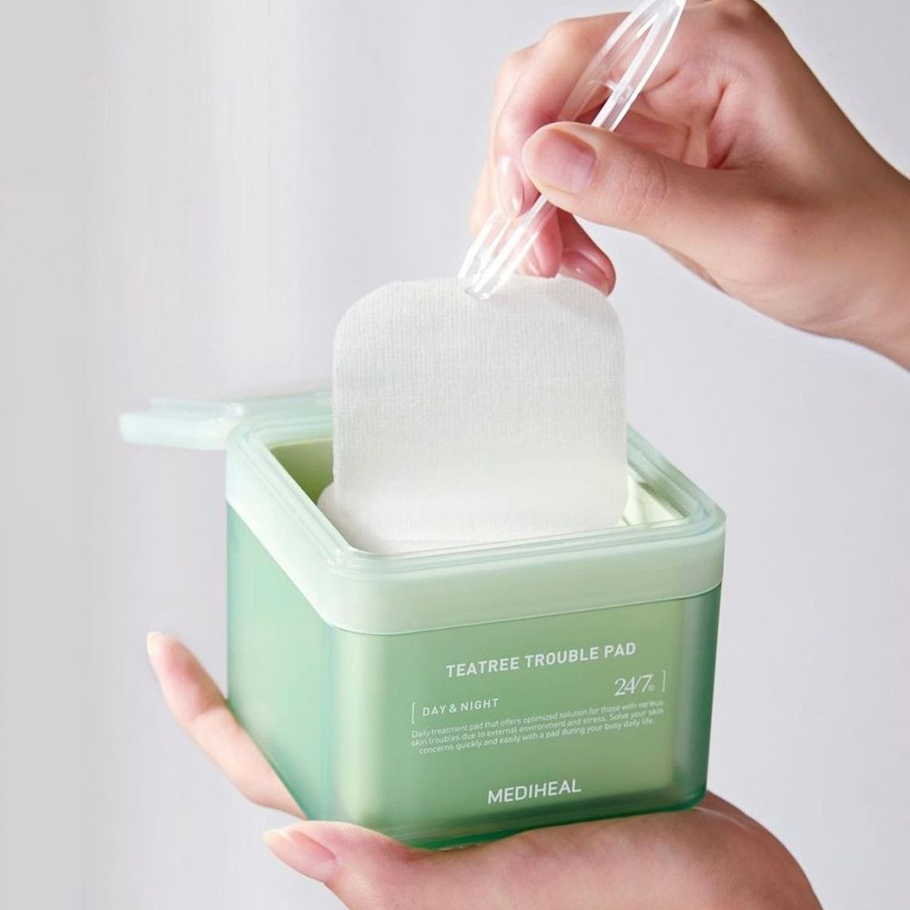 Scatola verde con dischetti. Una mano tiene la scatola, l'altra estrae un dischetto con una pinzetta. Scritta: TEA TREE TROUBLE PAD.