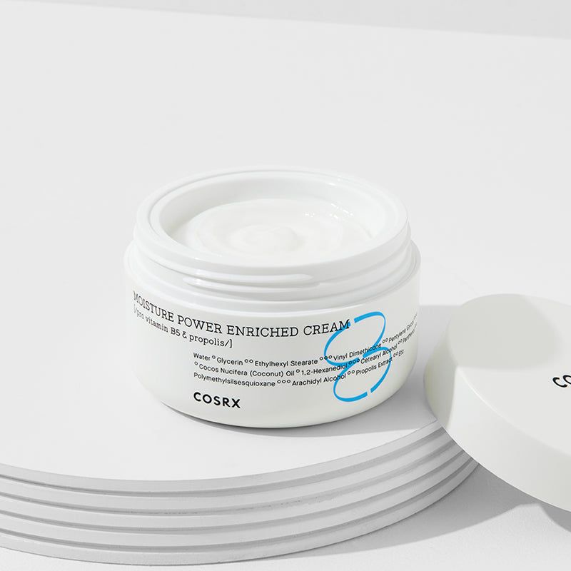 Vaso di crema bianco aperto su piedistallo bianco. Scritta: MOISTURE POWER ENRICHED CREAM, COSRX. Coperchio accanto.