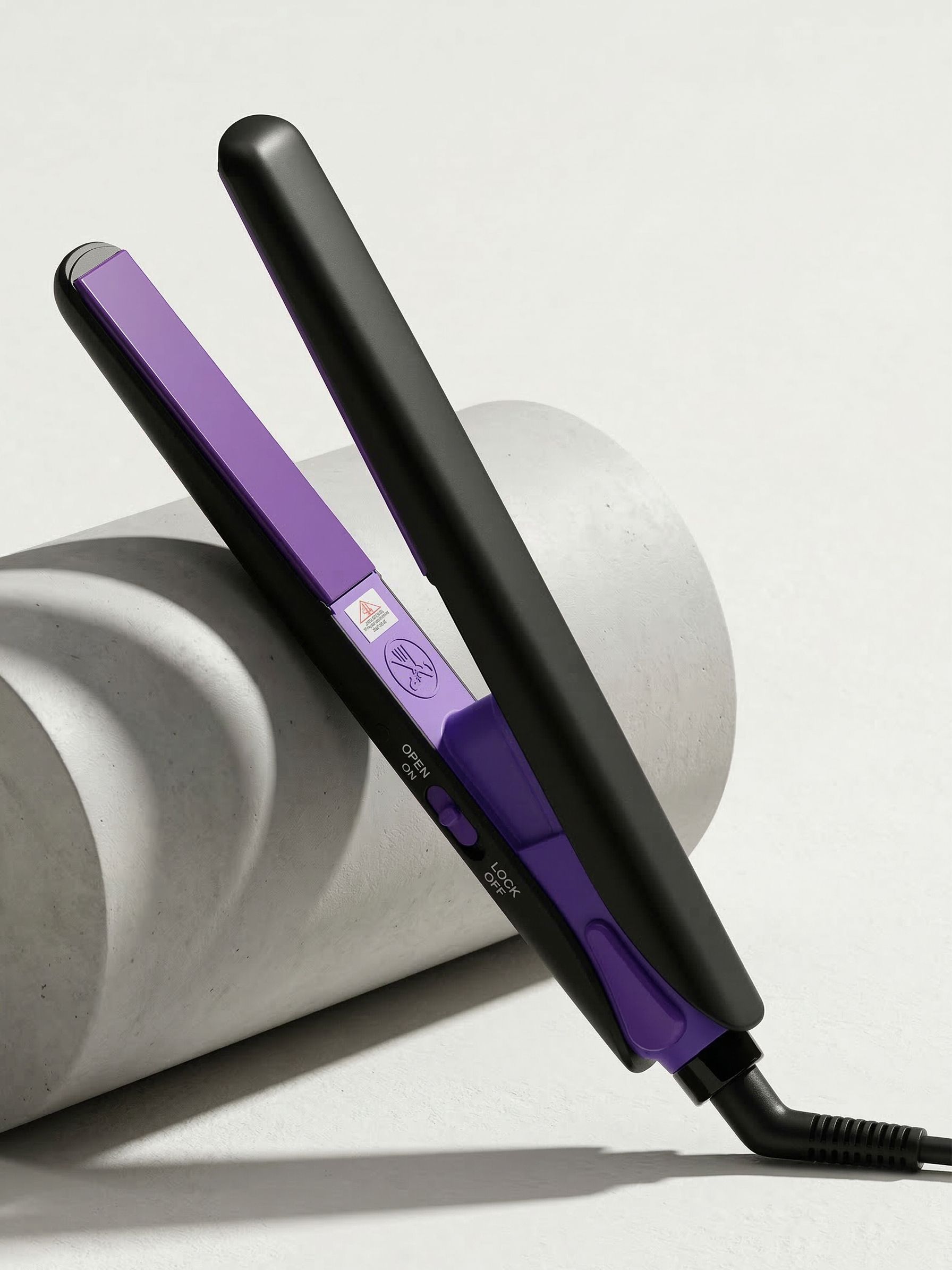 Mini piastra per capelli nera e viola. Piastre aperte, connessione via cavo. Su sfondo grigio.