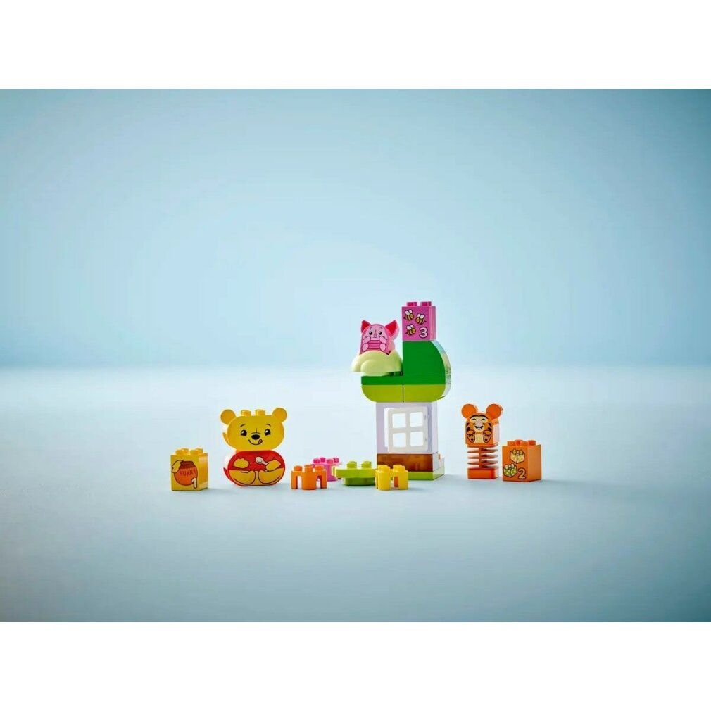 LEGO DUPLO 10457. Set con personaggi di Winnie the Pooh, casa, albero e accessori. Elementi singoli su sfondo blu.