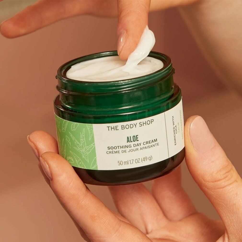 Mani che tengono un vasetto verde aperto con crema. Scritta: Aloe Soothing Day Cream. Crema in fase di prelievo.