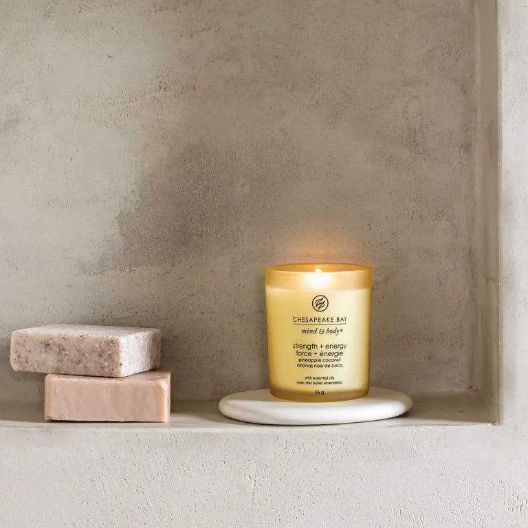 Candela gialla Chesapeake Bay mind & body, forza + energia, ananas cocco, su sottobicchiere bianco. Due saponi accanto.