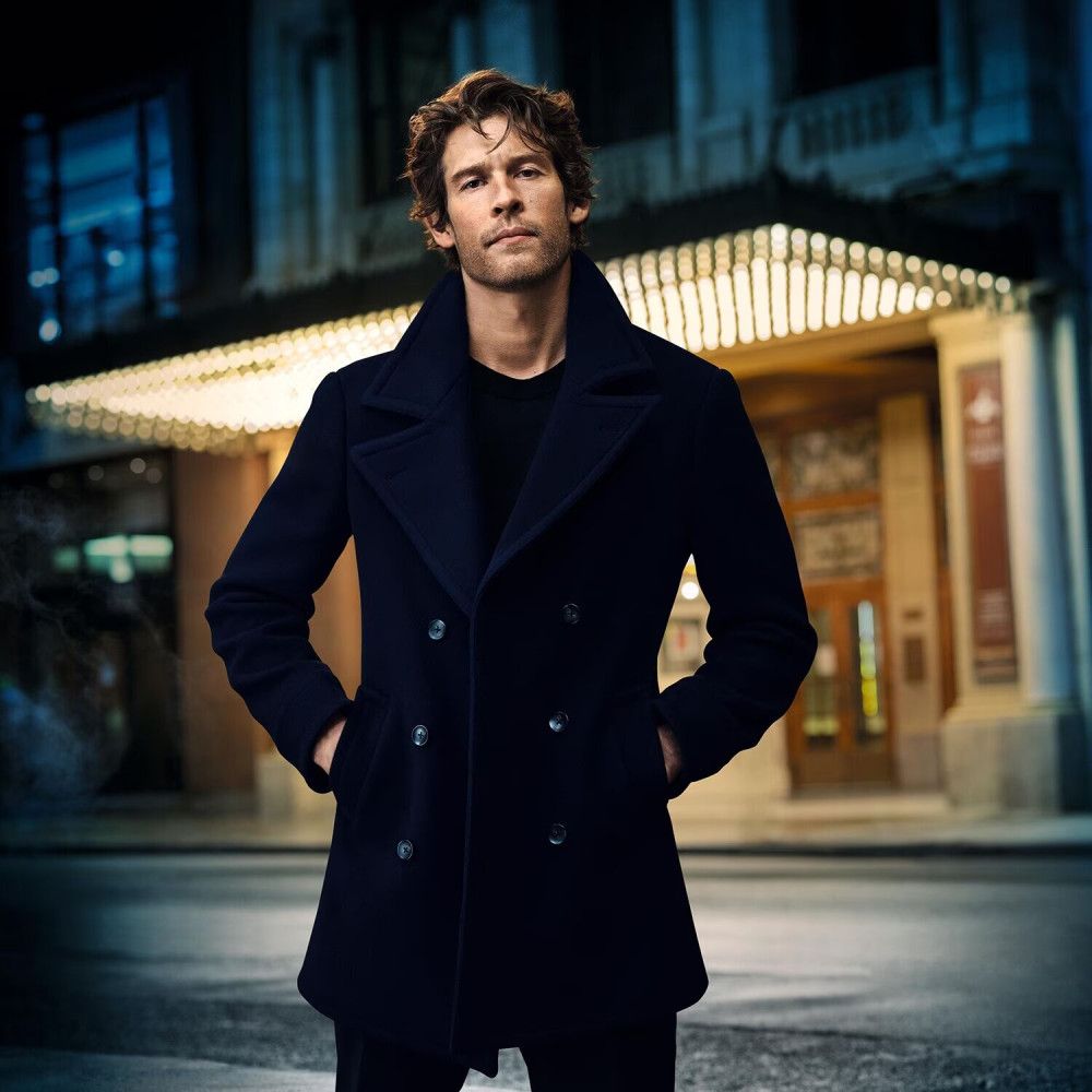 Uomo in cappotto blu scuro, davanti a un edificio illuminato. L'uomo è a braccia conserte.