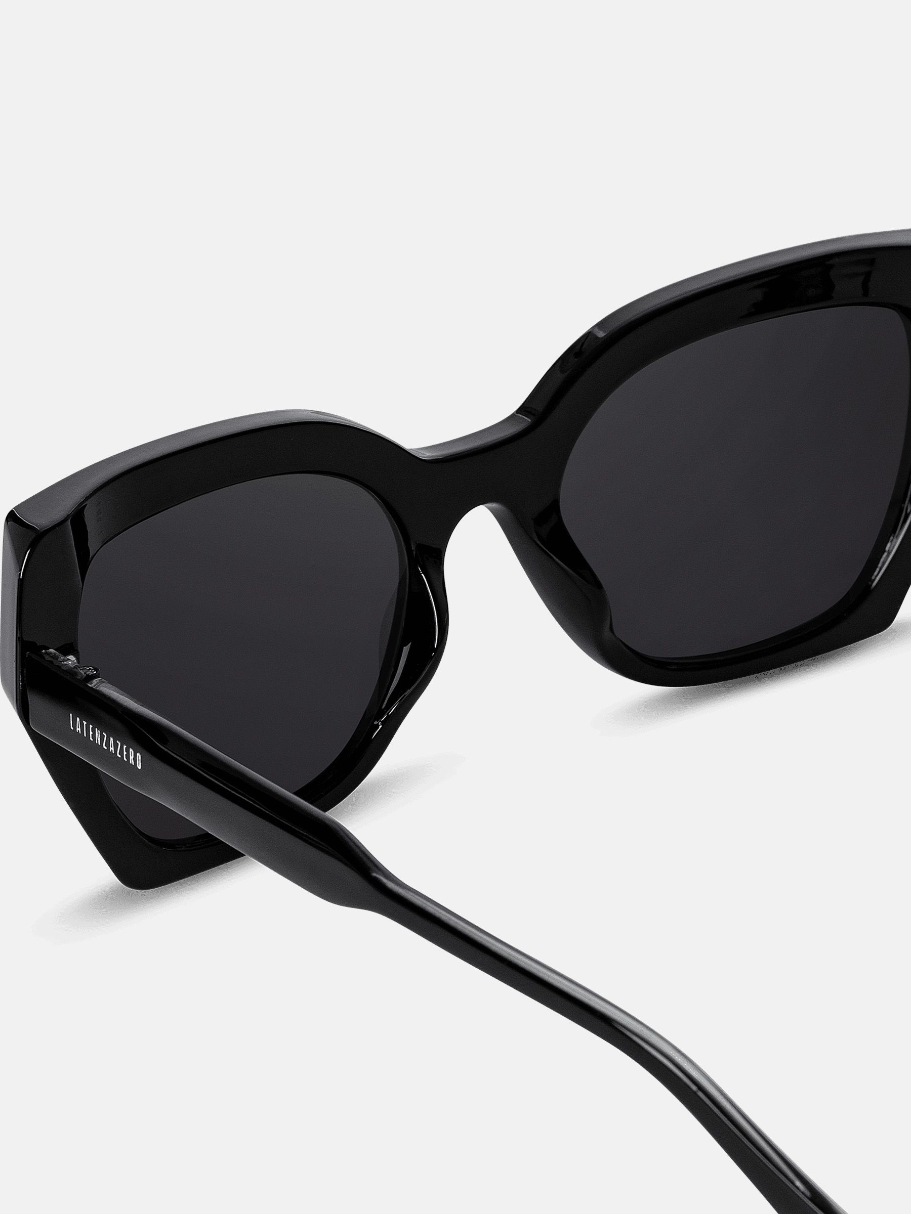 Primo piano di occhiali da sole neri. Lenti squadrate, montatura e aste nere. Prodotto leggermente inclinato.