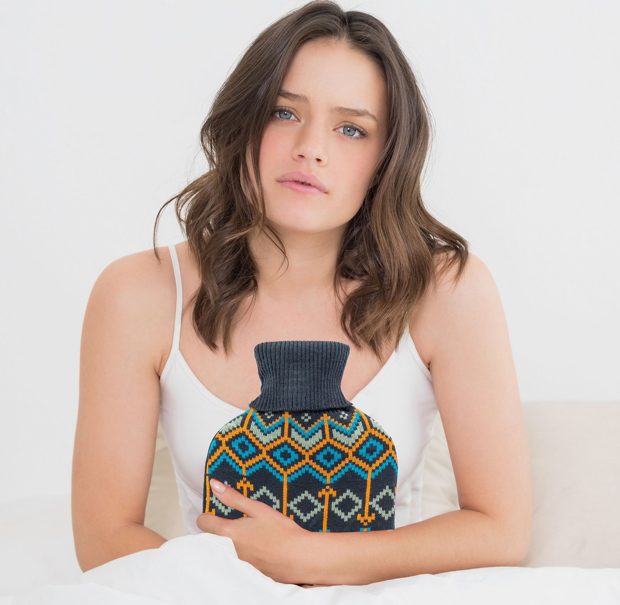 Donna seduta a letto, tiene borsa dell'acqua calda con copertura in maglia blu e gialla. Guarda la fotocamera.