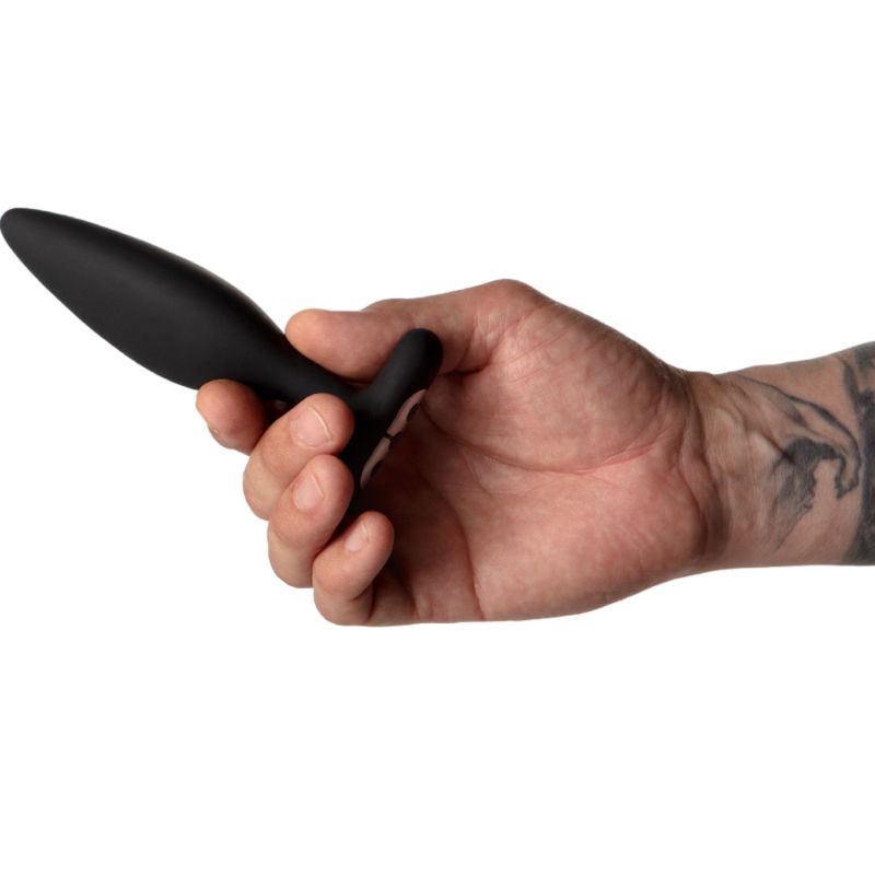 Mano che tiene un plug anale nero. Forma a T, punta arrotondata. Tatuaggio sul braccio.