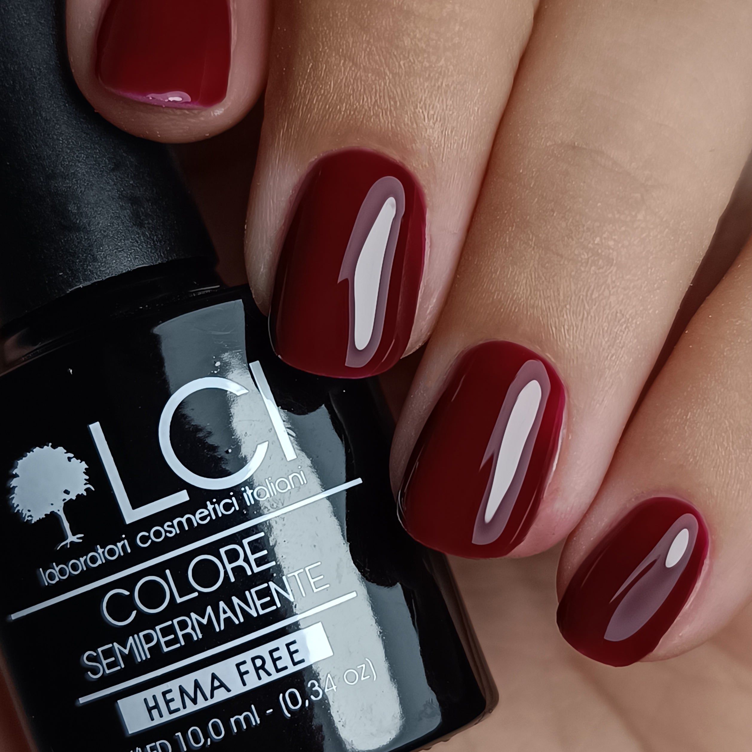 Mano con unghie smaltate e flacone di smalto. Smalto rosso scuro sulle unghie. Testo: LCI, COLORE SEMIPERMANENTE, HEMA FREE.