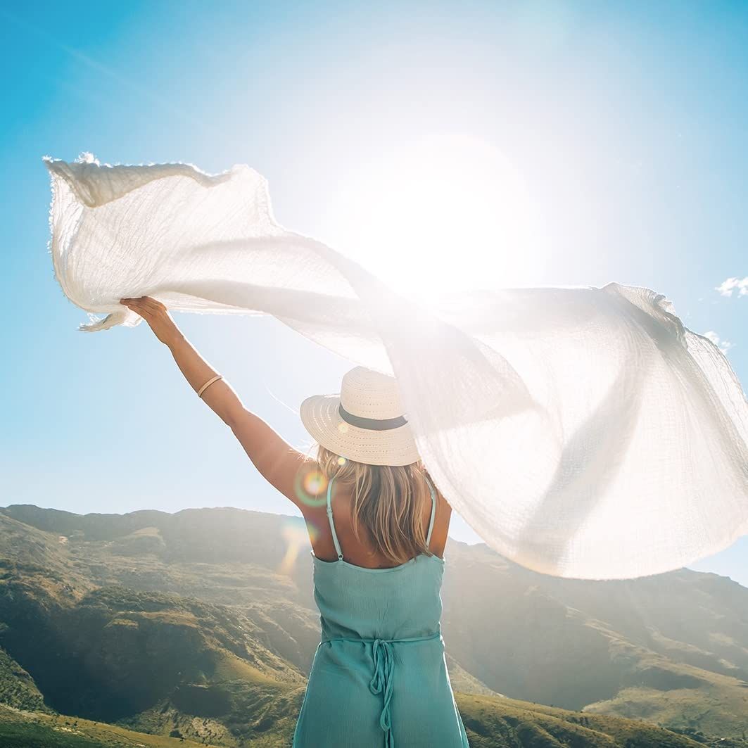 Donna con cappello e vestito, tiene un tessuto bianco al sole. Montagne sullo sfondo.