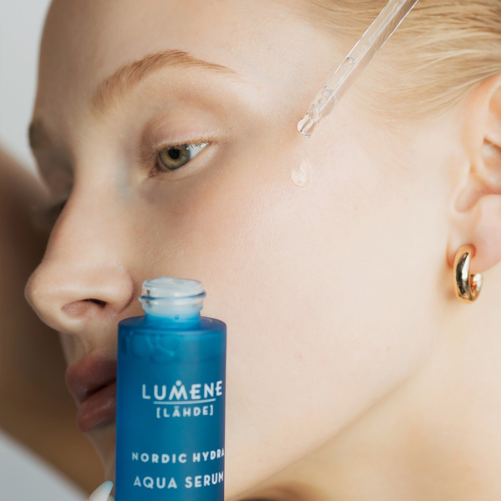 Donna con flacone e contagocce di siero. Il siero viene applicato sulla pelle. Scritta: Lumene [LAHDE] Nordic Hydra Aqua Serum.
