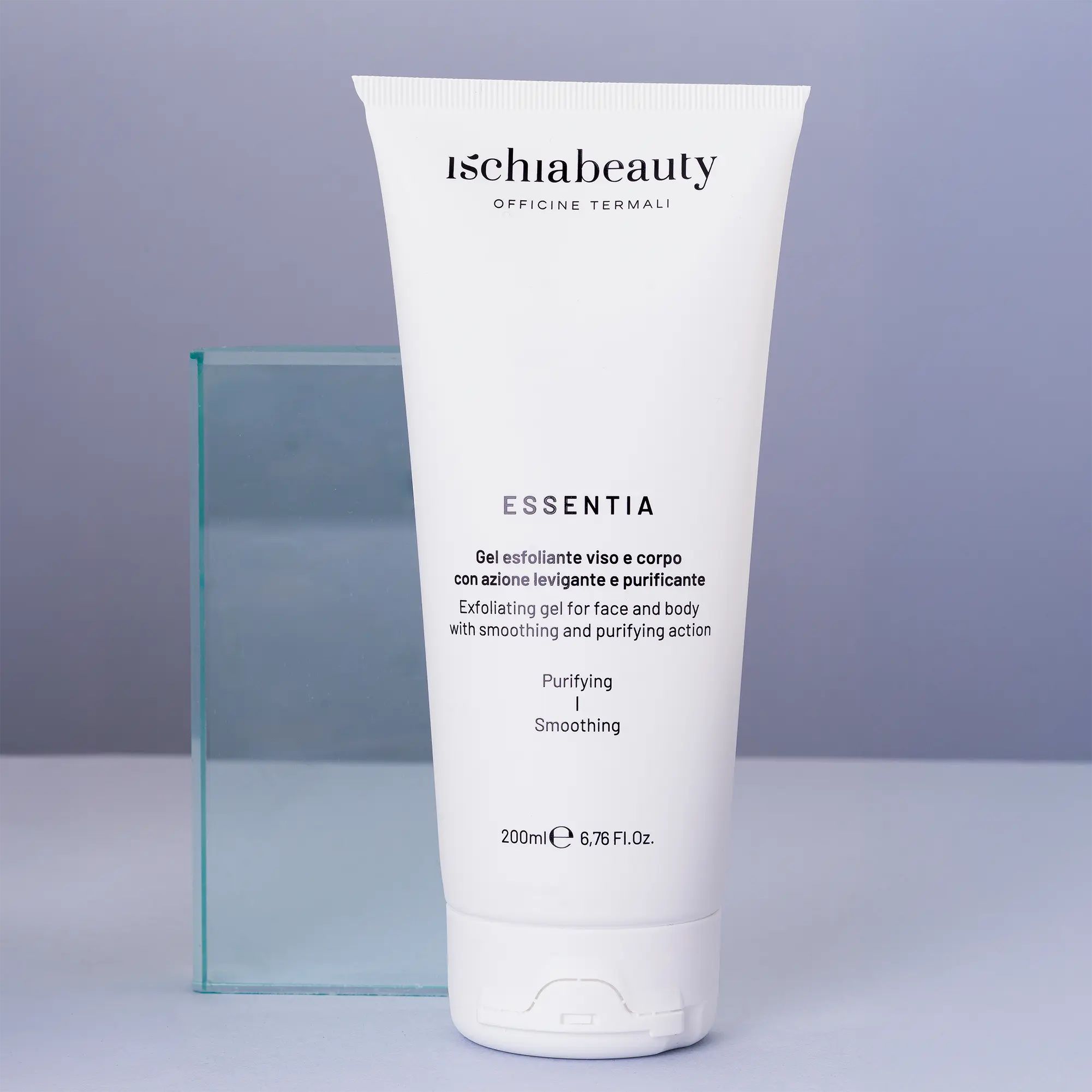 Tubo bianco con testo del prodotto. Testo: Ischiabeauty, Essentia, Gel esfoliante viso e corpo. Su sfondo blu, accanto a oggetto in vetro.